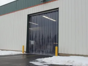 Flexible PVC strip curtains at a distribution center entrance, snow on the floor outside, early morning, the sky a pale grey